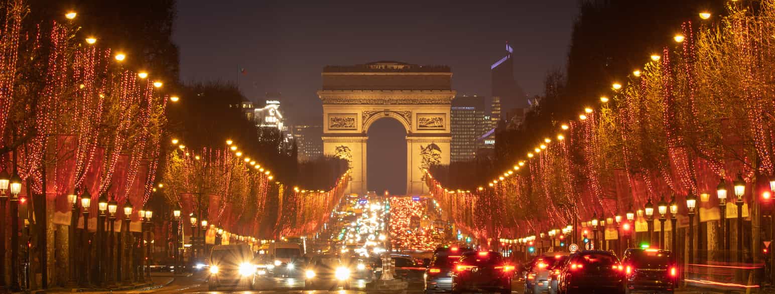 Champs-Élysées fotograferet om aftenen i retning mod triumfbuen. Alléens træer er pyntet med lyskæder, som lyser op, lige som bilernes lygter skaber lys i billedet.