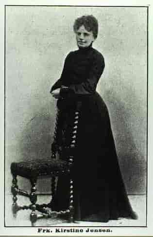 Gammelt sort-hvidt foto af en stående yngre kvinde, klædt i en hellang sort kjole,