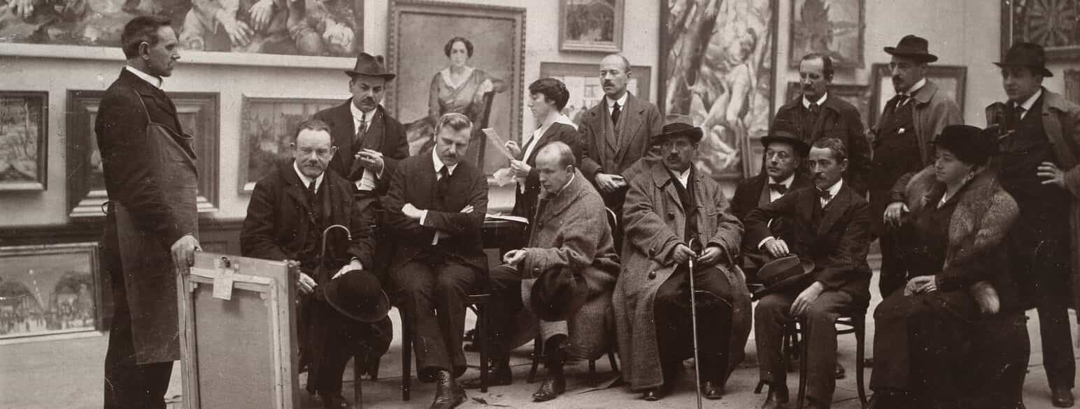 Medlemmer af Berliner Secession er samlet for at bedømme indsendte værker til en udstilling. I midten med hatten Lovis  Corinth og til venstre for ham Leo von König. Yderst til højre Charlotte Corinth. Foto fra 1905